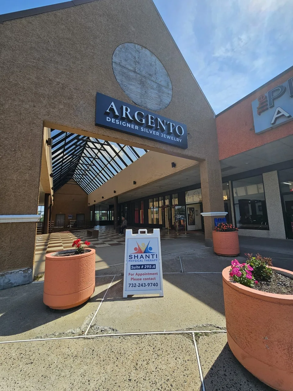 A white A-frame sign for Shanti Physical Therapy, Suite 295-A, in front of an arched plaza entrance.