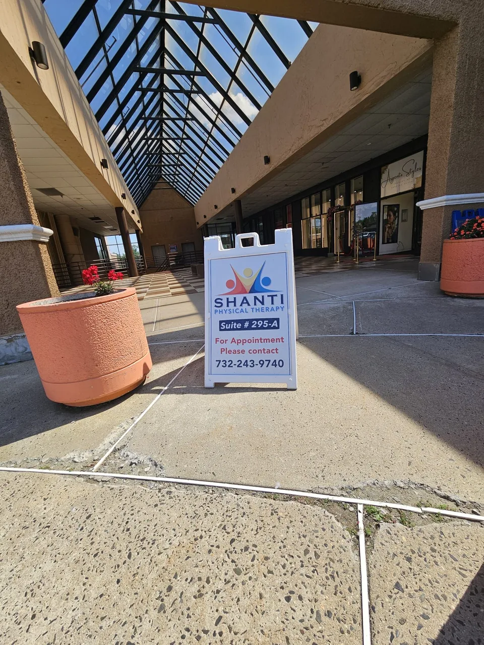 A white A-frame sign for Shanti Physical Therapy, Suite 295-A, standing in a covered plaza walkway with a glass roof.